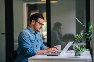 Serious pensive thoughtful focused young casual businessman or entrepreneur in office looking at and working with laptop making and typing serious important business email