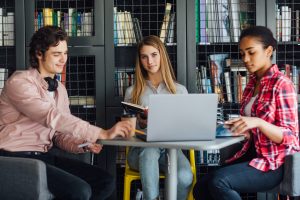 Three happy students writing to notebooks and laptop in library.