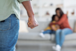 Domestic Violence. Unrecognizable African American Man Threatening Wife And Daughter With His Fist, Scared Mother Embracing Little Girl While Sitting Together On Couch, Selective Focus On Male Hand