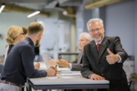 Senior businessman giving a thumbs-up sign while a group of other professionals are discussing something at a table.