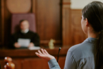 Young adult Asian woman testifying in courtroom, standing in front of judge, gesturing with hand while speaking, legal proceeding taking place in judicial setting