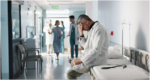 A mature male doctor in a white lab coat is sitting on a hospital bed in a hallway