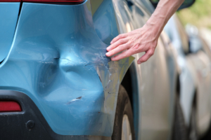 Driver hand examining dented car with damaged fender parked on city street side. Road safety and vehicle insurance concept.