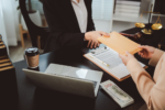 Two people exchange an envelope over a desk with contract, laptop, coffee, cash and courtroom symbols, suggesting a secretive legal or financial deal, possible bribery or fraud