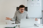 Person sitting at a desk with a laptop and raising their hands in a gesture of confusion or frustration