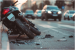 motorcycle lying on its side next to a curb on a paved street, surrounded by small debris, with blurred cars driving in the background