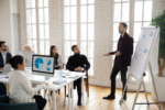 Full length young male team leader standing near whiteboard, presenting marketing research results to focused diverse investors at meeting. Concentrated smart business people listen to colleague.