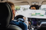 The image shows a driver inside a vehicle, viewed from behind the front passenger seat, focusing on the steering wheel, dashboard console, and a rear view mirror reflection