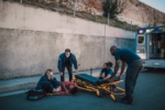 Emergency medical responders and a bystander attending to an individual lying on the pavement next to an open ambulance