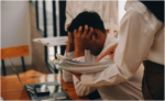 person sitting at a desk with their head in their hands while another person stands nearby