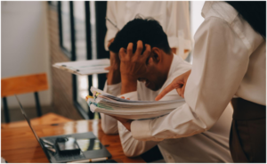 person sitting at a desk with their head in their hands while another person stands nearby