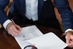 Person in a business suit signing a legal document at a dark wooden desk