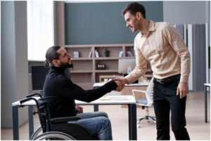 person in a wheelchair shaking hands with a standing colleague in an office setting