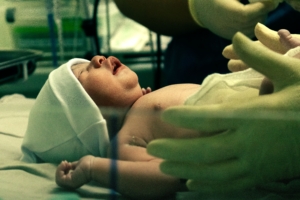 Newborn baby wearing a white cap lying on a hospital surface while being gently handled by gloved hands