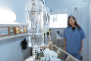 Intravenous (IV) infusion setup with a clear fluid bag hanging from a pole and a drip chamber in the foreground, and a medical worker in blue scrubs standing in the background of a clinical room